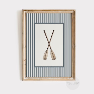 Puede incluir: Impresión artística enmarcada con dos remos de madera cruzados. La obra de arte está sobre un fondo blanco con un borde a rayas azul marino y blanco y un marco dorado. La impresión tiene un tema náutico.