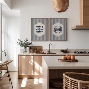 May include: Two framed nautical prints adorn a kitchen wall. One depicts fish, the other a crab, both on a blue and white striped backdrop. The kitchen features light wood cabinets, a white countertop, and a woven pendant light, creating a coastal theme.