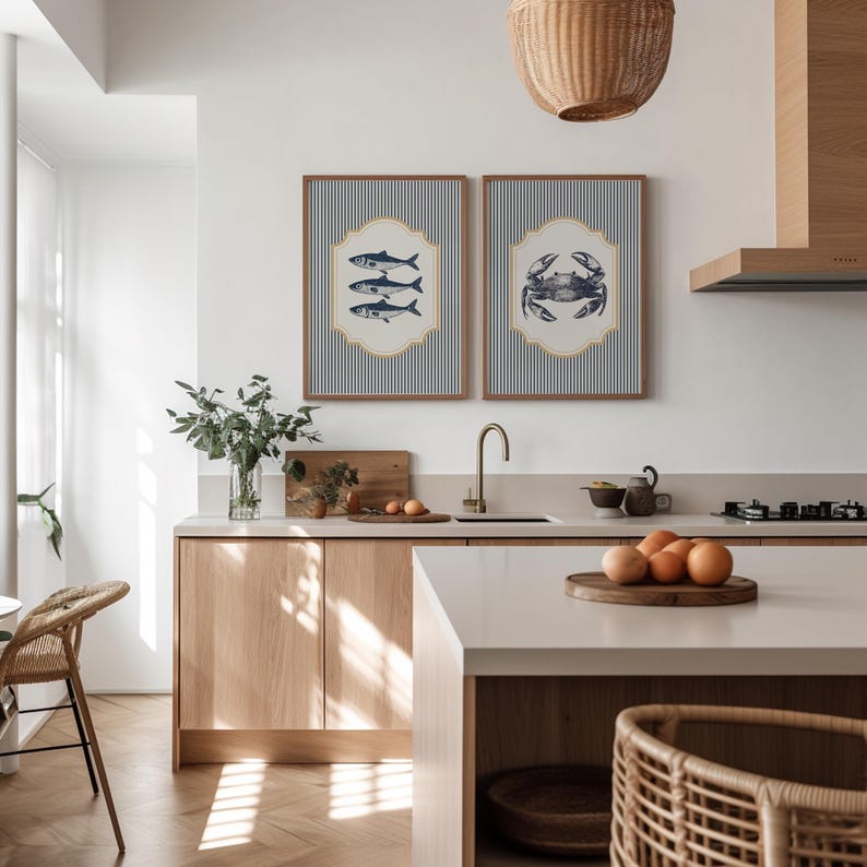 May include: Two framed nautical prints adorn a kitchen wall. One depicts fish, the other a crab, both on a blue and white striped backdrop. The kitchen features light wood cabinets, a white countertop, and a woven pendant light, creating a coastal theme.