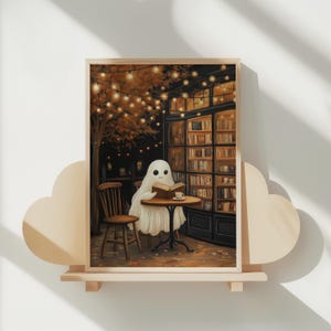 May include: A framed print depicts a friendly ghost reading a book at a cafe table. The scene is set at night, with warm lighting from string lights and a bookstore in the background. A wooden chair sits beside the table.