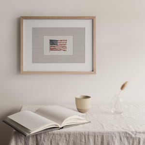 Peut inclure: Un cadre en bois clair encadre une impression du drapeau am&eacute;ricain. En dessous, un livre ouvert, une tasse en c&eacute;ramique et un petit vase en verre avec une plante s&eacute;ch&eacute;e sont pos&eacute;s sur une table recouverte de lin.