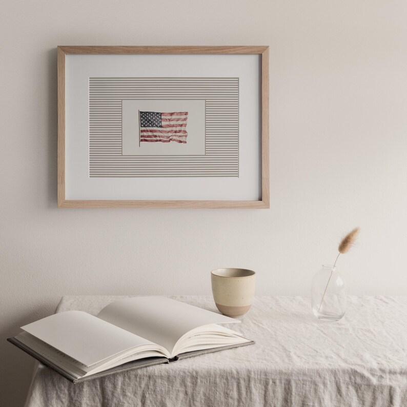 Peut inclure: Un cadre en bois clair encadre une impression du drapeau am&eacute;ricain. En dessous, un livre ouvert, une tasse en c&eacute;ramique et un petit vase en verre avec une plante s&eacute;ch&eacute;e sont pos&eacute;s sur une table recouverte de lin.