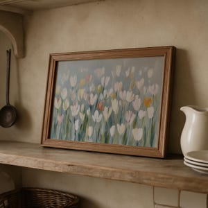 Puede incluir: Un cuadro rectangular enmarcado que presenta una escena floral impresionista con flores similares a tulipanes en blanco, rosa p&aacute;lido y amarillo suave sobre un fondo gris azulado, en un marco de madera de tono medio.