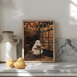May include: A framed print depicts a friendly ghost reading a book at a cafe table. The scene is set at night, with warm string lights and a bookstore in the background. The ghost is draped in a white sheet, and a cup of coffee sits on the table.