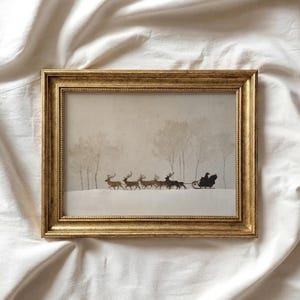 May include: A rectangular gold-toned frame containing a print of a silhouette scene. The artwork depicts a dark figure driving a sleigh pulled by a team of reindeer across a snowy landscape with bare trees in the background, all in muted tones.
