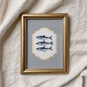 May include: A gold-framed print displaying three blue fish illustrations on a white and gold decorative shape, set against a blue and white striped background. The frame is rectangular.
