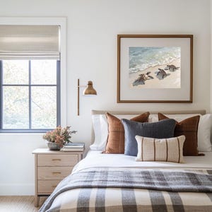 Op de afbeelding: Een slaapkamer met een wit bed, een bruin-wit gestreept dekbed en een ingelijst afdruk van zeeschildpadden op het strand. Het bed heeft verschillende kussens in bruin, wit en blauw.