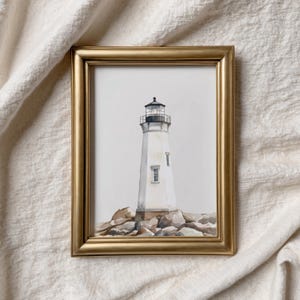 May include: A framed watercolor painting of a white lighthouse with a black top, set on a rocky base. The lighthouse has a small window and is surrounded by gray rocks. The frame is gold.
