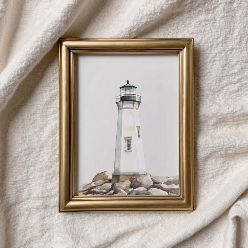 May include: A framed watercolor painting of a white lighthouse with a black top, set on a rocky base. The lighthouse has a small window and is surrounded by gray rocks. The frame is gold.