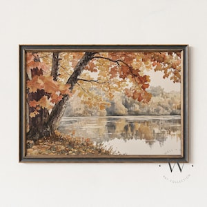 Op de afbeelding: Aquarel schilderij van een serene herfstscène aan het meer. Het kunstwerk toont een grote boom met oranje en gele bladeren, die weerspiegelen in het kalme water. Het schilderij is ingelijst in een donkere houten lijst.
