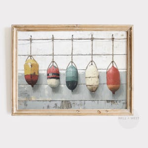 May include: A framed print showing five colourful buoys suspended by ropes against a weathered, painted wooden backdrop. The buoys are painted in various colours, including yellow, red, white, and teal, with a distressed, vintage aesthetic. The frame is a light wood colour.