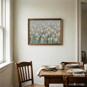 Puede incluir: Un cuadro rectangular enmarcado que presenta un campo impresionista de tulipanes blancos, amarillo p&aacute;lido y rosa suave con tallos verdes sobre un fondo gris azulado, expuesto en una pared sobre una mesa de comedor de madera con sillas.