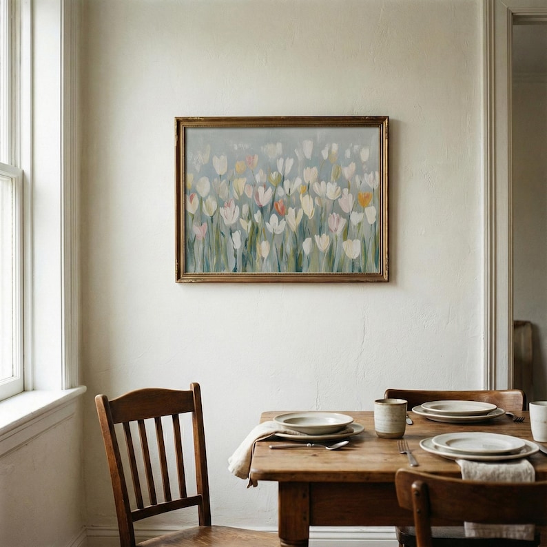 Puede incluir: Un cuadro rectangular enmarcado que presenta un campo impresionista de tulipanes blancos, amarillo p&aacute;lido y rosa suave con tallos verdes sobre un fondo gris azulado, expuesto en una pared sobre una mesa de comedor de madera con sillas.