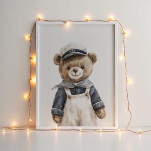 May include: A framed watercolor illustration of a brown teddy bear wearing a white sailor hat with an anchor emblem, a blue and white striped neckerchief, and white overalls over a dark blue long-sleeved shirt, surrounded by string lights.