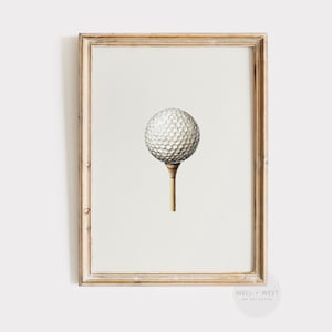May include: A framed print showing a golf ball on a tee. The golf ball is white with a dimpled texture, and the tee is light brown. The frame is a light wood colour, and the background is a neutral off-white. A classic golf-themed artwork.