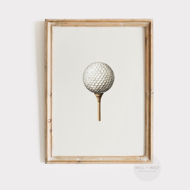 May include: A framed print featuring a detailed illustration of a golf ball on a tee. The golf ball is white with a dimpled surface, and the tee is a light brown color. The frame is a light wood tone, and the background is a neutral off-white.