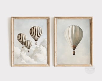 May include: Two framed prints of hot air balloons. The print on the left shows three hot air balloons in the sky with clouds. The print on the right shows one hot air balloon in the sky with clouds. Both prints have a light brown frame.