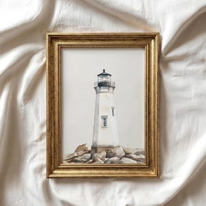 May include: A framed watercolor painting of a white lighthouse with a black top, set on a rocky base. The lighthouse has a small window and is framed in a gold-colored, ornate frame. The artwork is displayed against a white background.