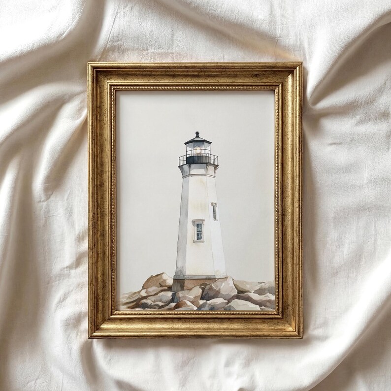 May include: A framed watercolor painting of a white lighthouse with a black top, set on a rocky base. The lighthouse has a small window and is framed in a gold-colored, ornate frame. The artwork is displayed against a white background.