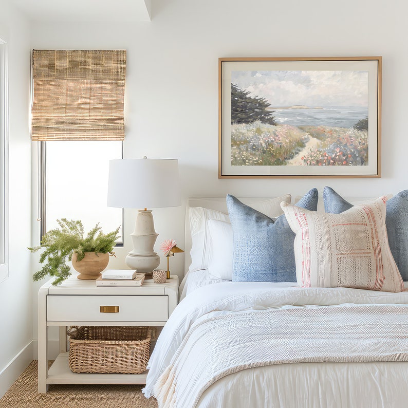 May include: A bedroom scene featuring a white bed with blue pillows and a decorative pillow with a pink and white pattern. A framed landscape painting hangs above the bed. A white nightstand holds a lamp, a plant, and a decorative basket.
