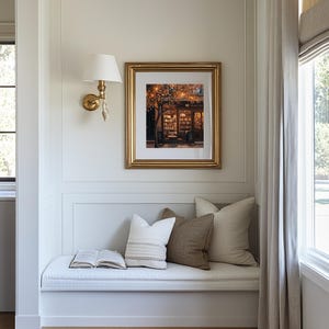 May include: A framed print with a gold frame depicts a cozy bookstore scene with warm lighting. The print hangs above a window seat with pillows and an open book. A gold sconce with a white shade is mounted on the wall.