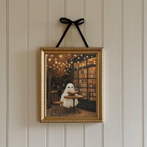 May include: A framed art print featuring a whimsical ghost reading a book at a cafe table. The artwork has a gold frame and is suspended by a black ribbon. The scene includes a cafe setting with a bookshelf, string lights, and a wooden chair.