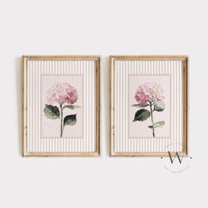 Può includere: Due stampe artistiche floreali incorniciate. Ogni stampa presenta un fiore di ortensia rosa con foglie verdi su uno sfondo a righe rosa e bianche. Le cornici sono di colore legno chiaro e l'arte ha uno stile acquerello.