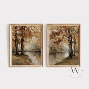 May include: Two framed landscape paintings depict a serene autumn scene. The artwork features trees with brown and gold leaves reflected in a calm body of water. The frames are a light brown color.