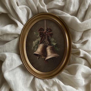 May include: An oval, gold-framed artwork featuring two bronze bells, a red velvet bow, and evergreen sprigs. The artwork is set against a dark background and hangs from a thin string. The frame is displayed on a white, textured fabric.