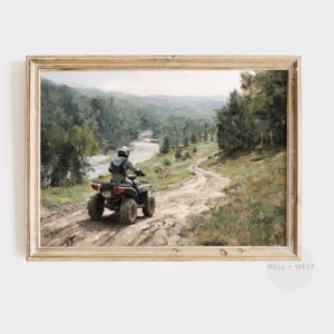 May include: A framed landscape print shows a person on an ATV on a dirt track through a forest. The artwork features a river winding through the valley, with lush green trees and a light sky. The print is in a gold-coloured frame.