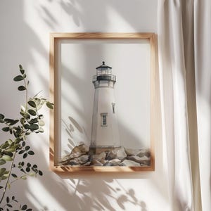 May include: Watercolor print of a white lighthouse with a black top, set against a white background. The lighthouse sits on a rocky base, framed in a light wood. The artwork is displayed on a white wall with shadows from a nearby plant.