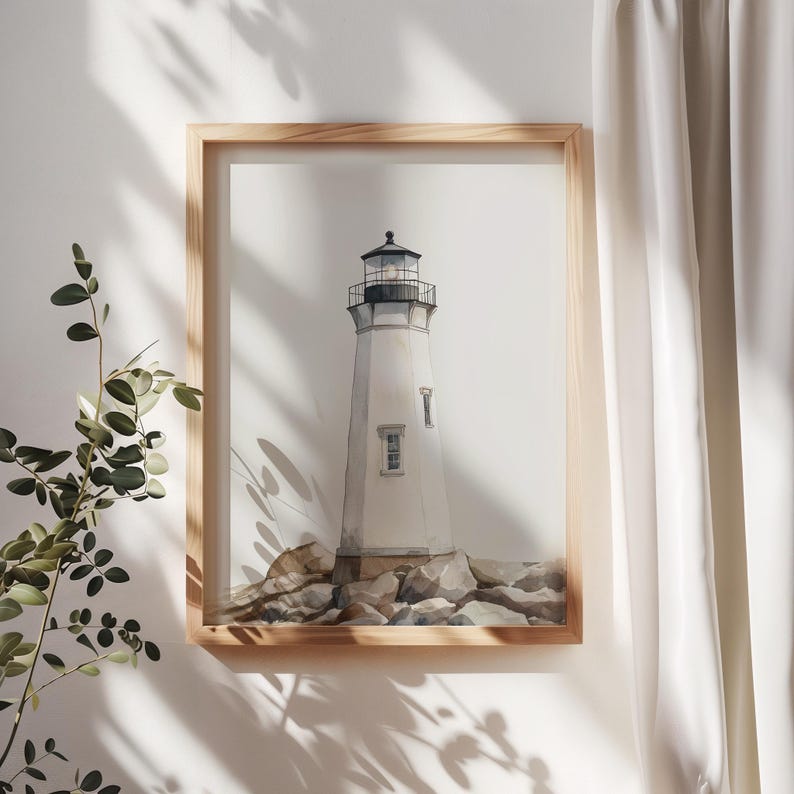 May include: Watercolor print of a white lighthouse with a black top, set against a white background. The lighthouse sits on a rocky base, framed in a light wood. The artwork is displayed on a white wall with shadows from a nearby plant.