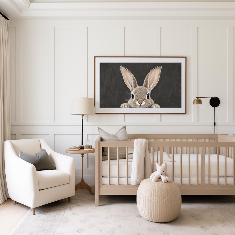 May include: A white crib with a white mattress and a white blanket. A brown rabbit is peeking over the top of a framed picture hanging on the wall. A white armchair with a gray pillow is in the foreground. A small round table with a lamp is next to the armchair.