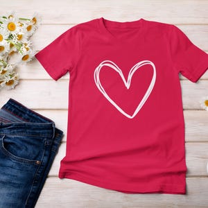 以下が含まれることがあります： 白い手書きのハートのデザインが施された赤いTシャツ。Tシャツは、青いジーンズ、白いデイジーの花束、青と白のスニーカーと一緒に木製の表面に置かれています。