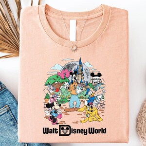 May include: A peach colored t-shirt with a graphic of Mickey Mouse and other Disney characters in front of Cinderella's Castle. The text "Walt Disney World" is printed below the graphic.