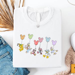 a white t-shirt with a colorful graphic of Disney characters, including Mickey Mouse, Donald Duck, Goofy, Minnie Mouse, and Donald Duck, along with balloons. The t-shirt is laid out on a white surface, along with a pair of blue jeans, white sneakers, and a beige blanket.