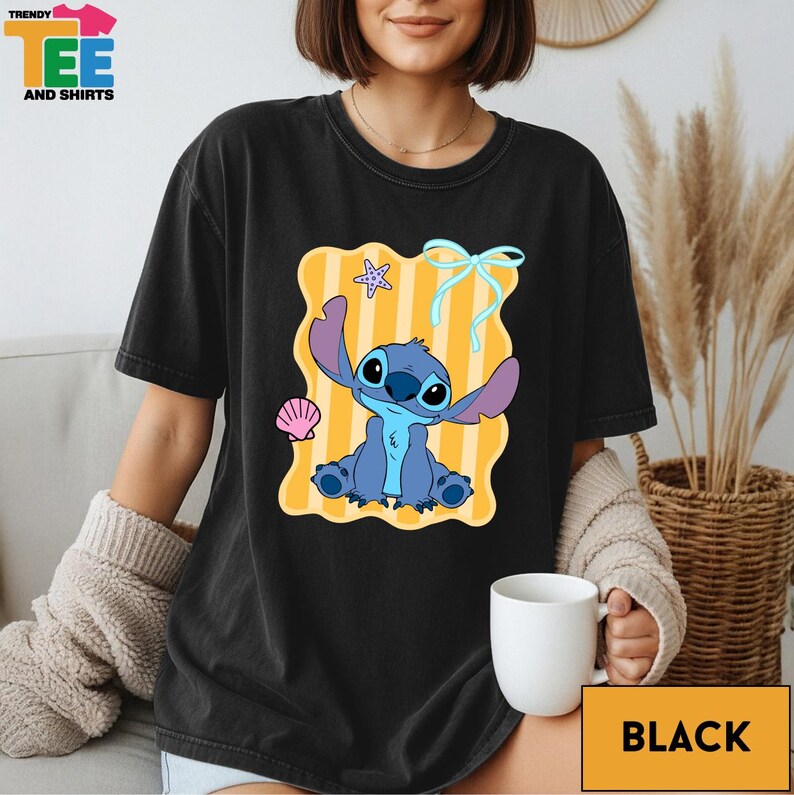 A woman wearing a black t-shirt with a colorful graphic of a cartoon character, specifically Stitch, sitting on a beach with a seashell and starfish.