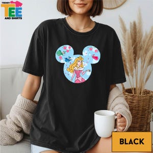 A woman wearing a black t-shirt with a Disney character design is sitting on a couch, holding a white mug.