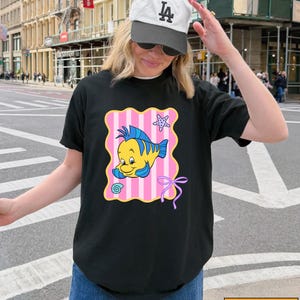 A woman wearing a black t-shirt with a cartoon fish graphic and a white cap is standing on a city street.
