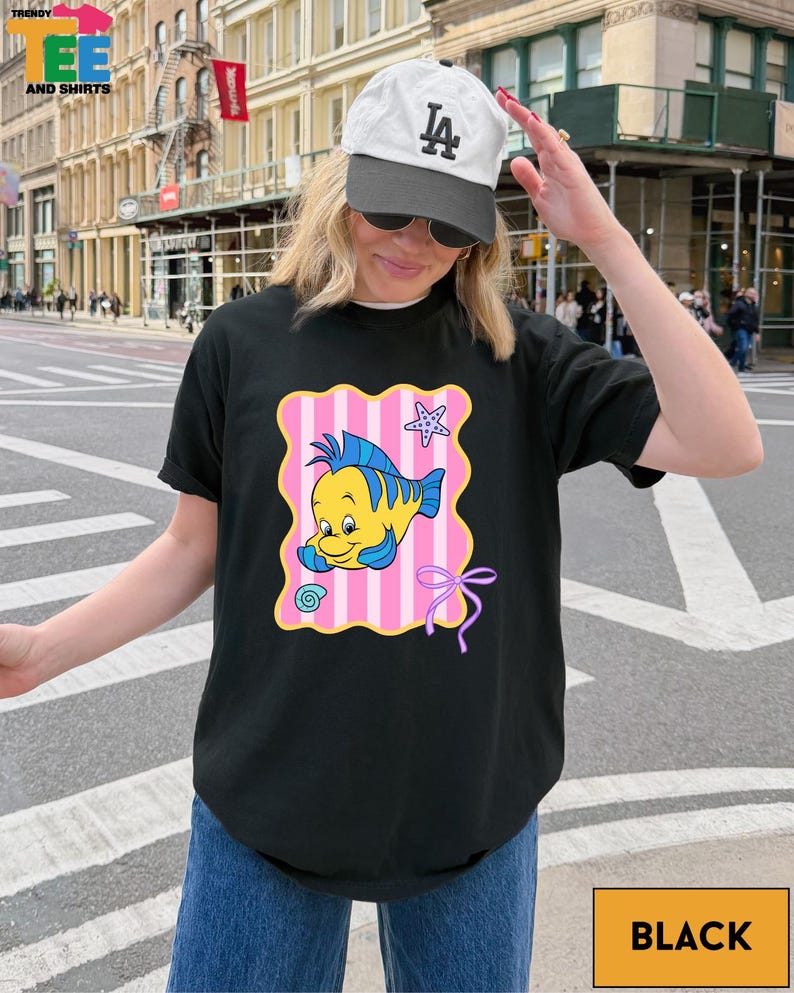 A woman wearing a black t-shirt with a cartoon fish graphic and a white cap is standing on a city street.