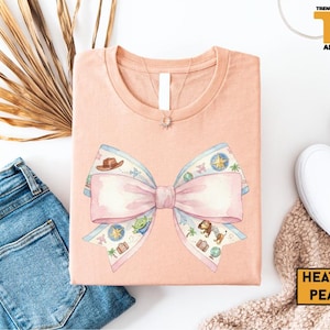 a pink t-shirt with a bow design, a pair of blue jeans, a pair of white sneakers, and some decorative elements such as a palm leaf and a beige blanket.