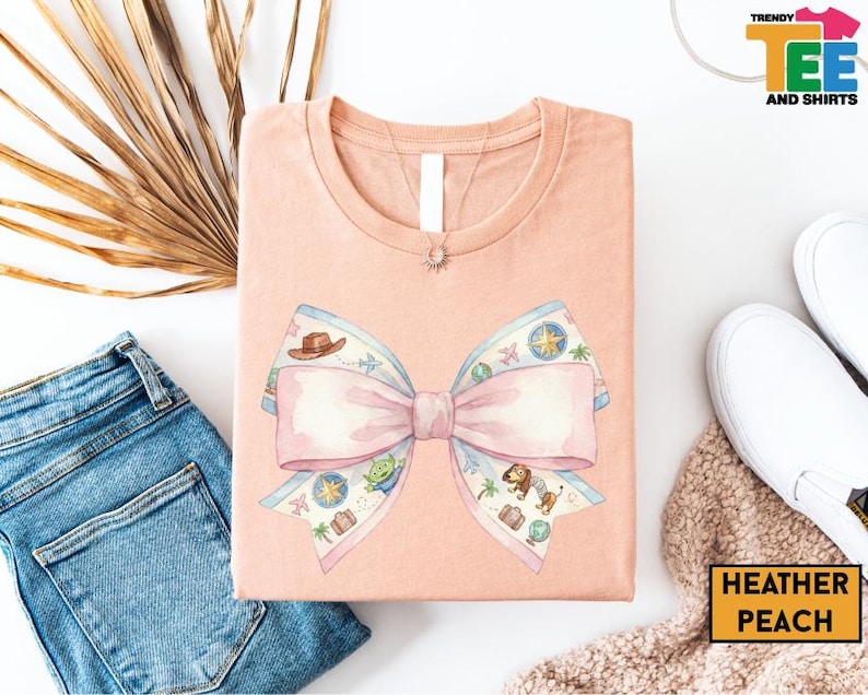 a pink t-shirt with a bow design, a pair of blue jeans, a pair of white sneakers, and some decorative elements such as a palm leaf and a beige blanket.