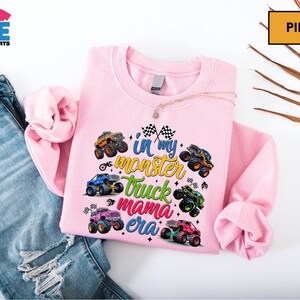 A pink sweatshirt with a colorful design of monster trucks and the text "I'm in my monster truck era" printed on it, along with a necklace. The sweatshirt is placed on top of a pair of blue jeans, and there are some leaves in the background.