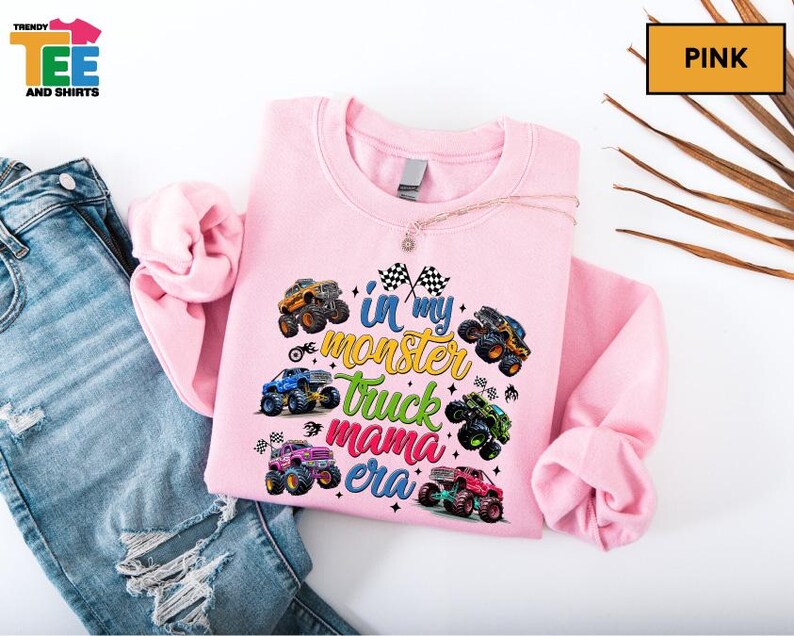 A pink sweatshirt with a colorful design of monster trucks and the text "I'm in my monster truck era" printed on it, along with a necklace. The sweatshirt is placed on top of a pair of blue jeans, and there are some leaves in the background.