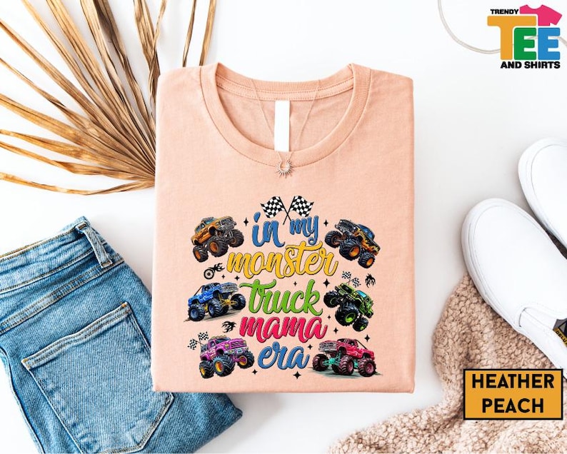 a pink t-shirt with a graphic design of monster trucks, along with a pair of jeans and a pair of white sneakers.