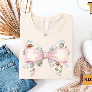 a casual outfit consisting of a white t-shirt with a pink bow, a pair of blue jeans, and a pair of white sneakers, all laid out on a white background.