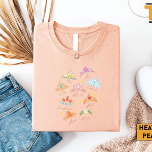 a casual outfit consisting of a pink t-shirt with a crown design, a pair of blue jeans, white sneakers, and a beige sweater.