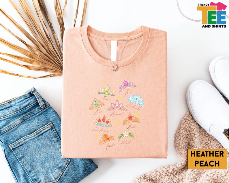 a casual outfit consisting of a pink t-shirt with a crown design, a pair of blue jeans, white sneakers, and a beige sweater.