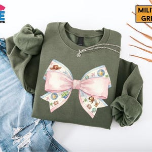 a green sweatshirt with a large pink bow on it, along with a pair of blue jeans and some leaves.