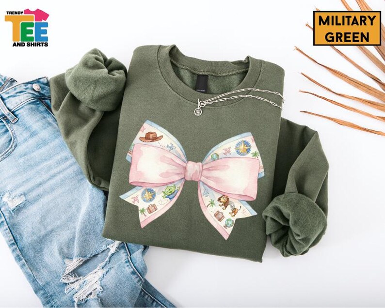 a green sweatshirt with a large pink bow on it, along with a pair of blue jeans and some leaves.